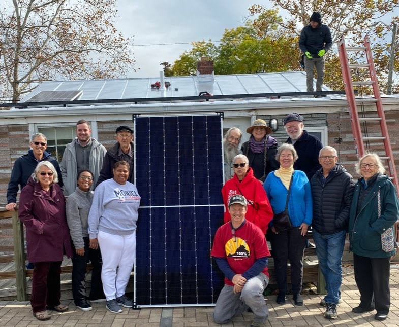Community members gathered around a solar-panel installation, with solar panels visible on the roof behind them.