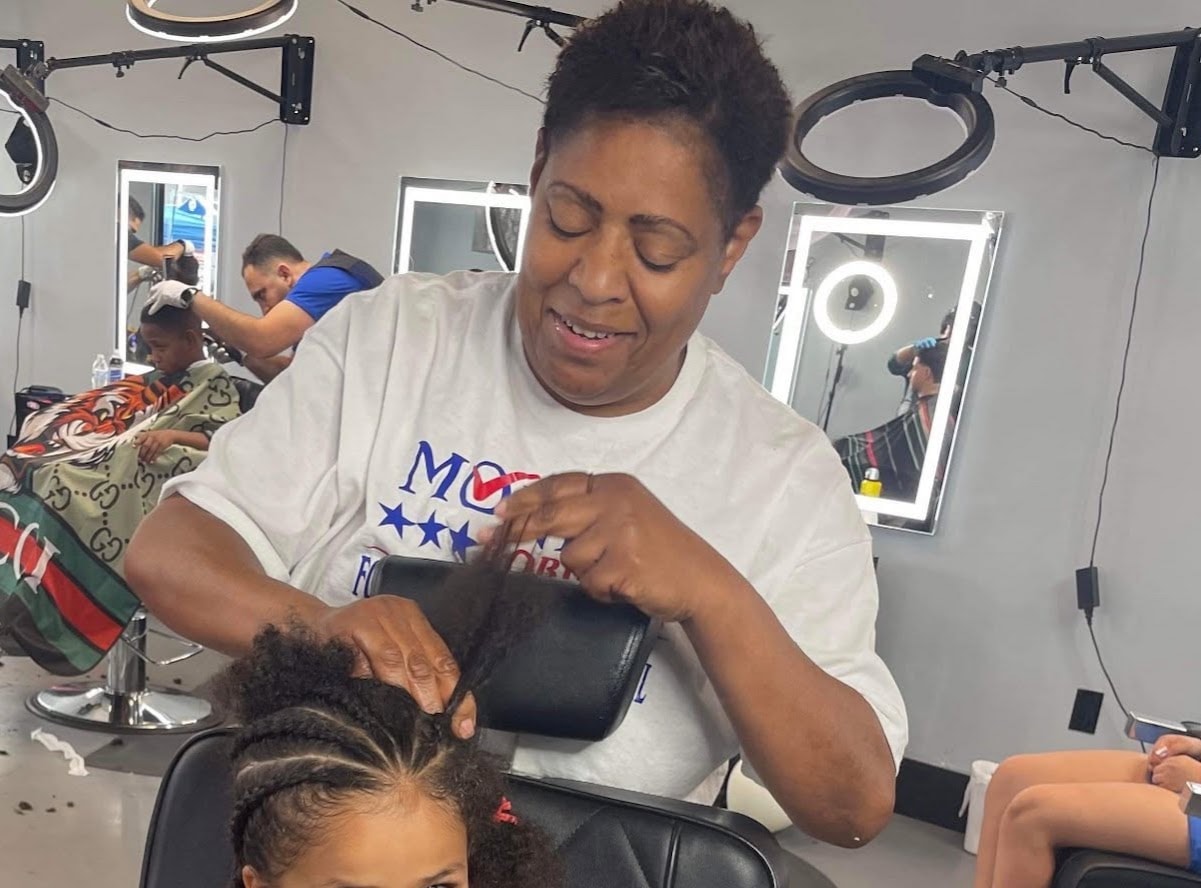 Monica Robinson braiding a young community member's hair in a salon, surrounded by other stylists and clients.